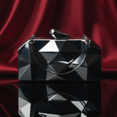 The Manay black geometric clutch purse with metallic finish on glossy surface, red drape backdrop