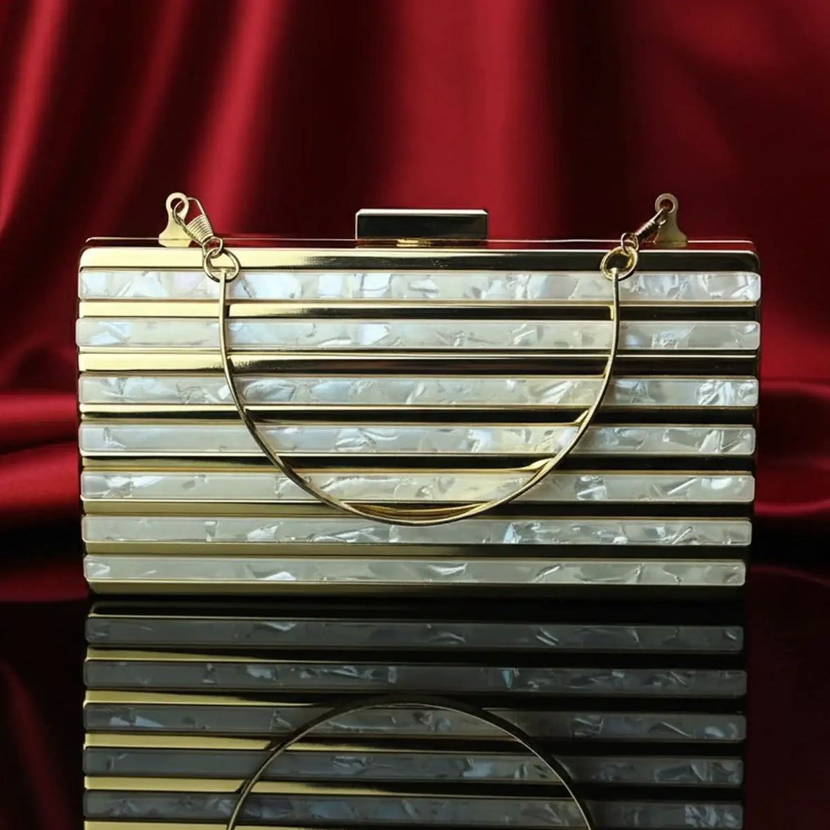 The Manay gold and pearl acrylic clutch bag on shiny black surface with red satin backdrop