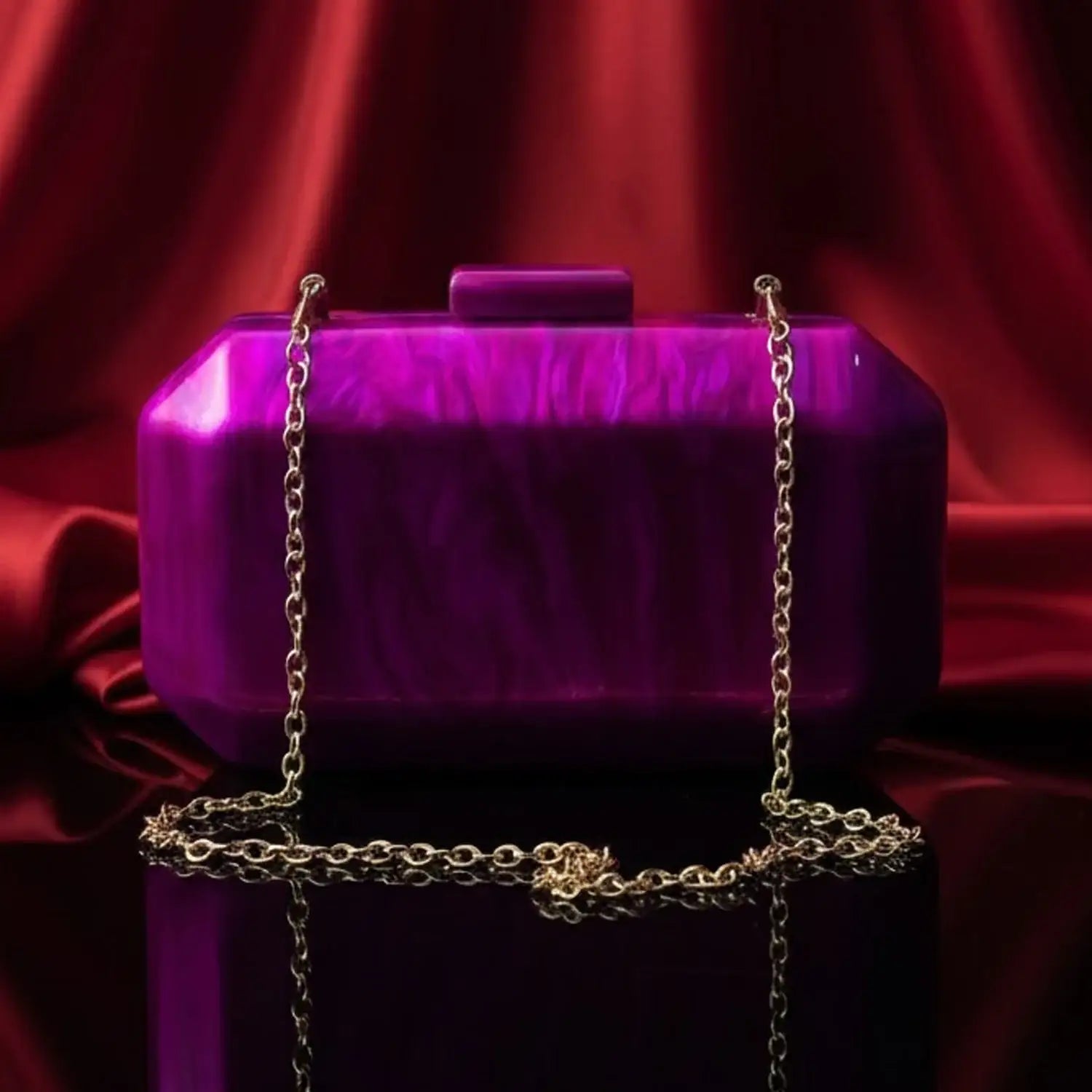 The Manay purple acrylic clutch bag with gold chain on glossy surface and red satin backdrop