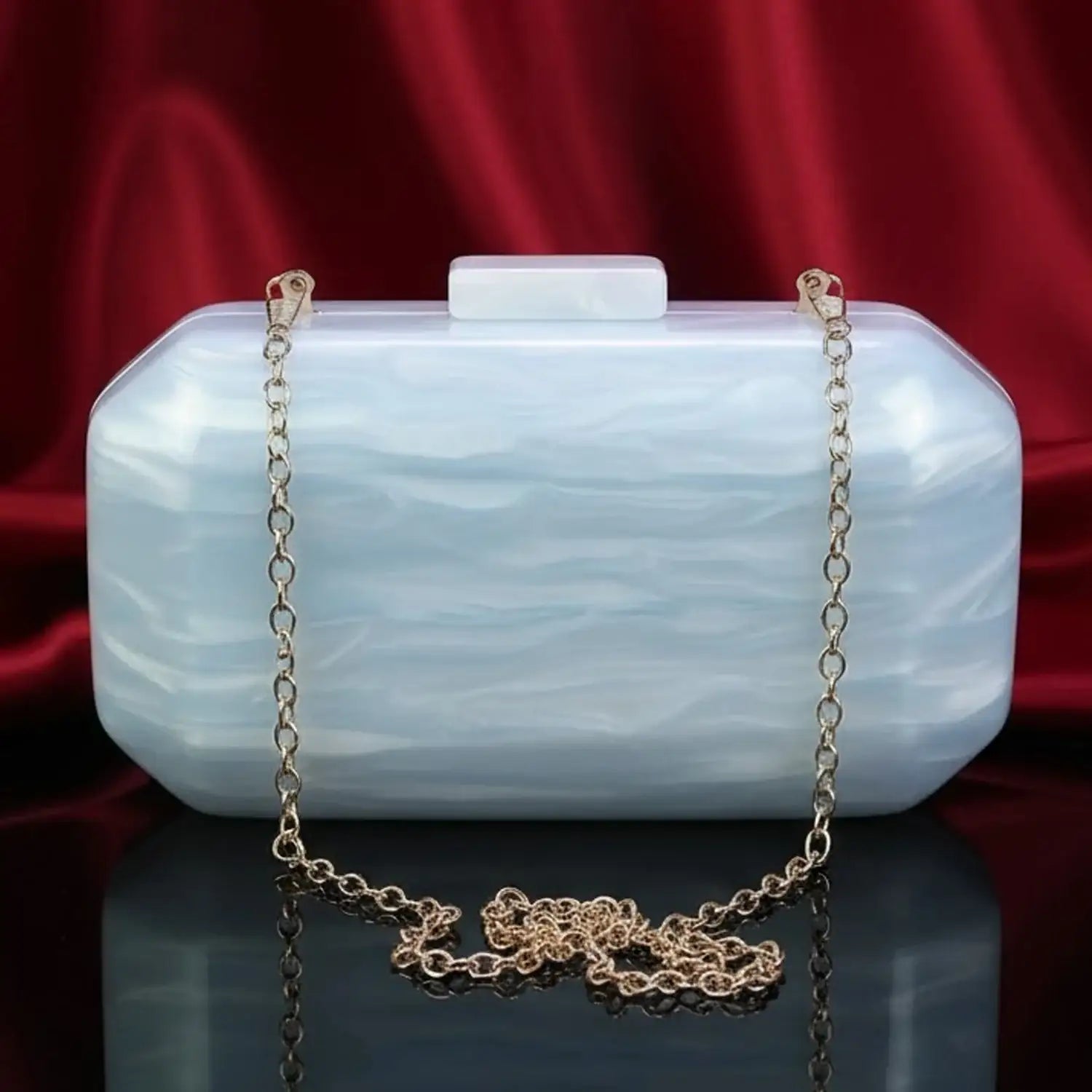 Elegant white acrylic clutch bag with gold chain on black surface, The Manay