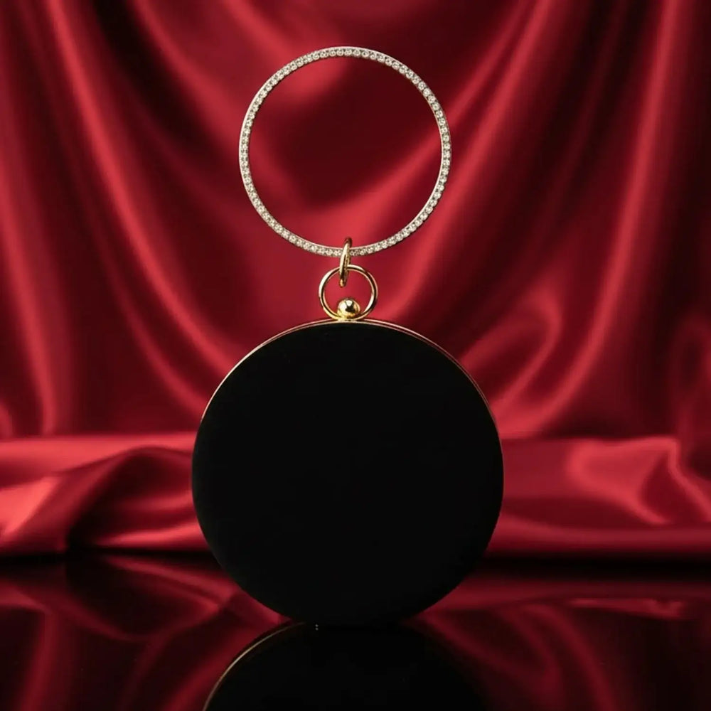 The Manay black round evening clutch with gold ring handle on red satin backdrop
