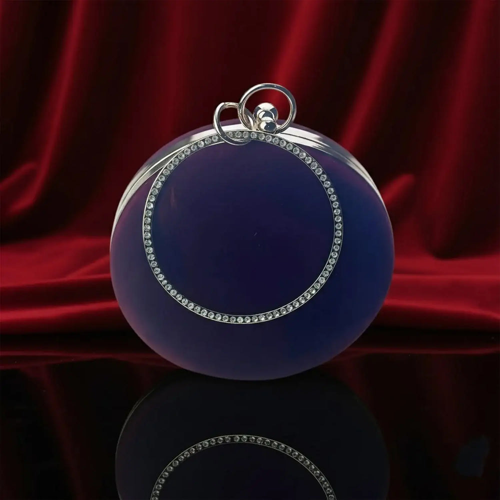 The Manay luxury round blue clutch with crystal trim on black surface, red velvet backdrop